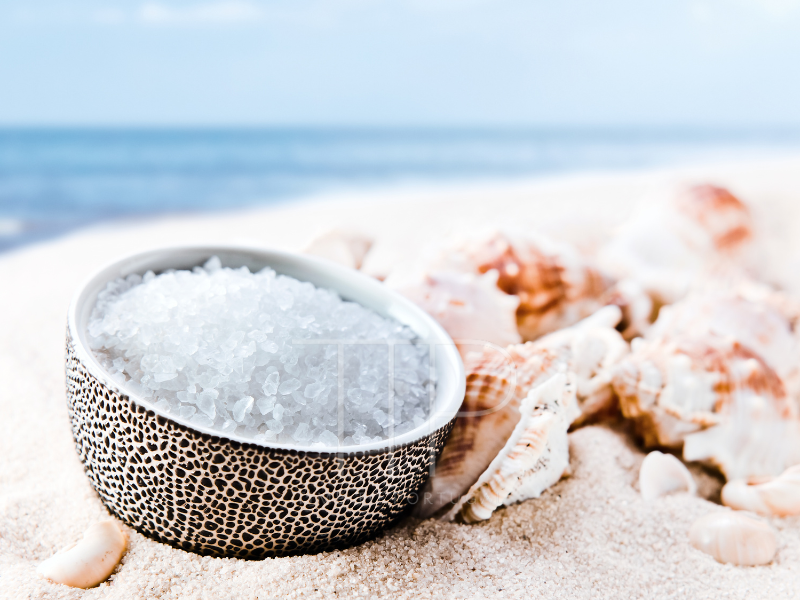 small bowl of sea salt besides sea shells on the beach