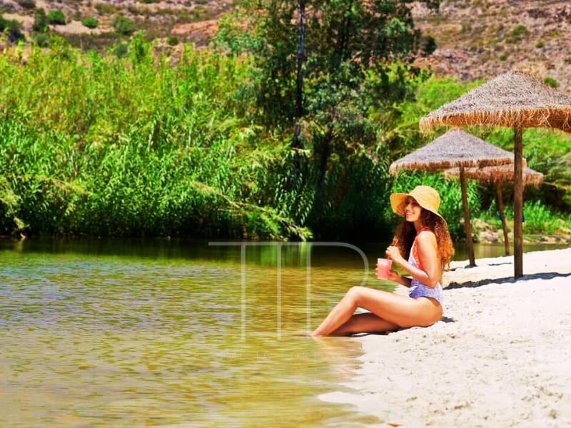A woman sitting on a river beach with feet in the water.